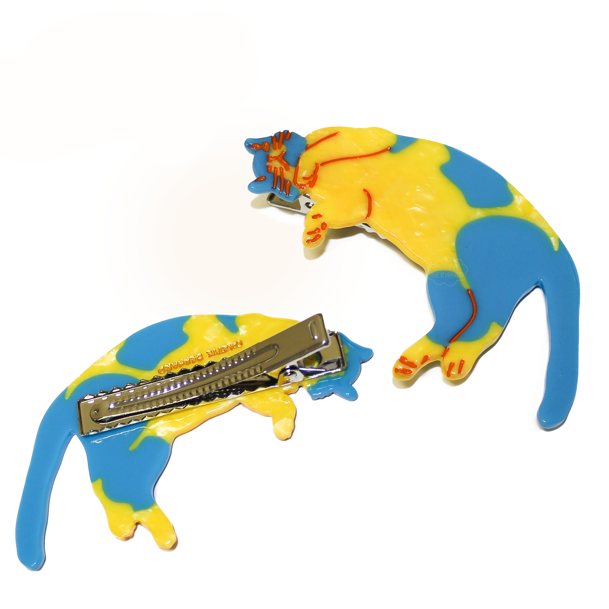 A hair clip shaped like a cat laying on its back. the cat is yellow with blue patches, and is outlined in red to show details. The image shows two clips, one flipped over showing the metal alligator clip mechanism. the other clip is facing up, showing the cat design