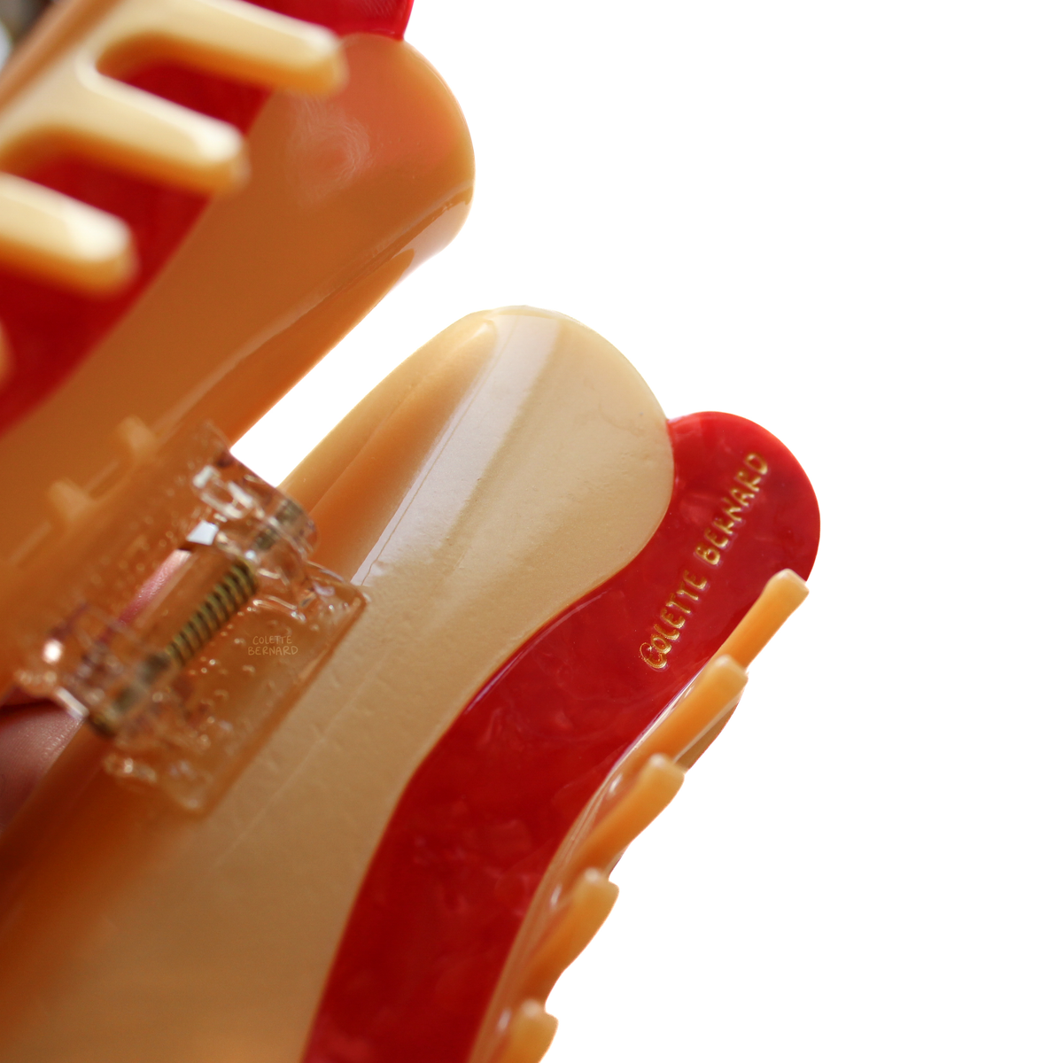 A close up of the Hot Dog Hair Claw held open, showing the spring mechanism, the beige teeth and the gold Colette Bernard logo on the red edge.
