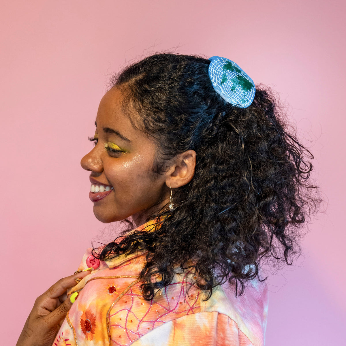 A person with dark curly hair wearing the World Map Hair Claw half up, shown from the side smiling against a pink background.