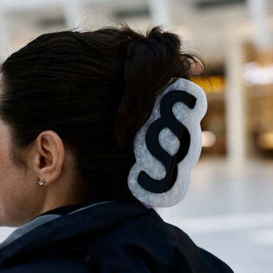 A person with dark hair wearing the Section Sign Mega Claw, a large white pearl claw with a black section sign symbol, in a bun, seen from behind in a blurred indoor space.