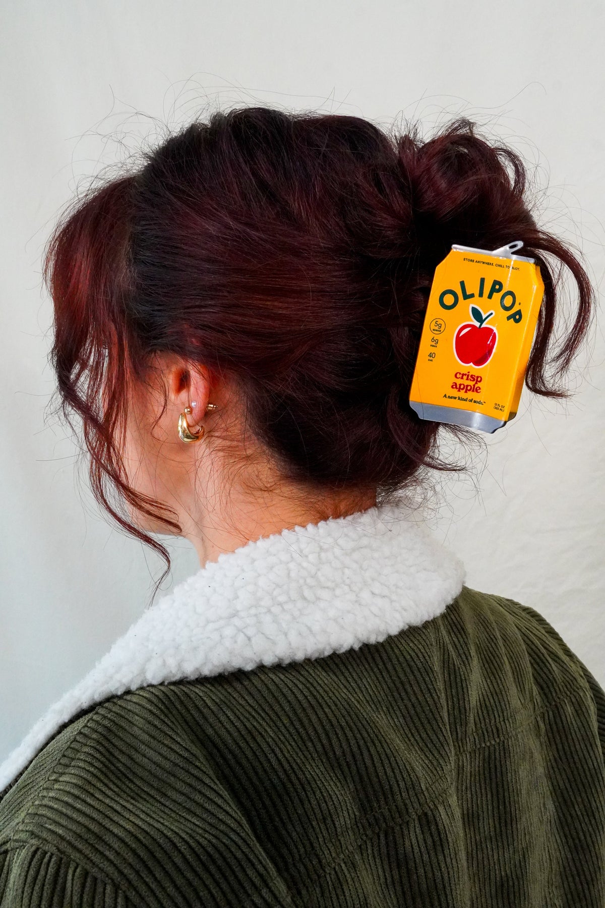 A person shown from the back wearing a dark olive green corduroy jacket with a white sherpa collar, with their hair in a bun secured by a OLIPOP Crisp Apple Hair Claw