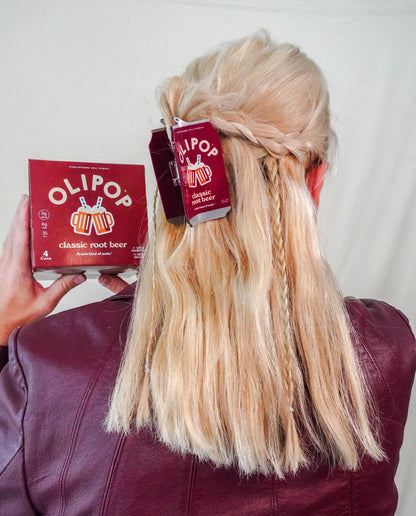 A person shown from behind wearing a maroon leather jacket, with long blonde hair styled in a half-up braid. They are holding a OLIPOP classic root beer box in their left hand, with a matching OLIPOP classic root beer hair claw holding the hair