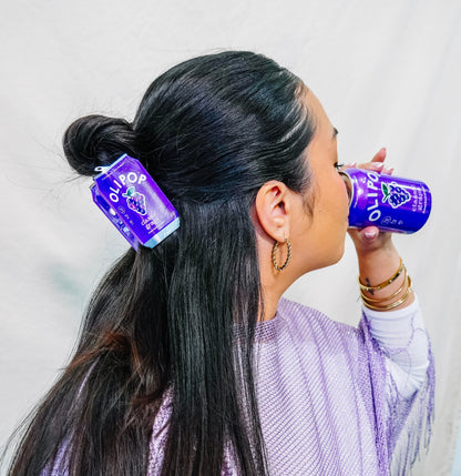 A woman with long dark hair half up shown in profile drinking from a Olipop Classic Grape can while wearing a matching  Olipop Classic Grape hair claw
