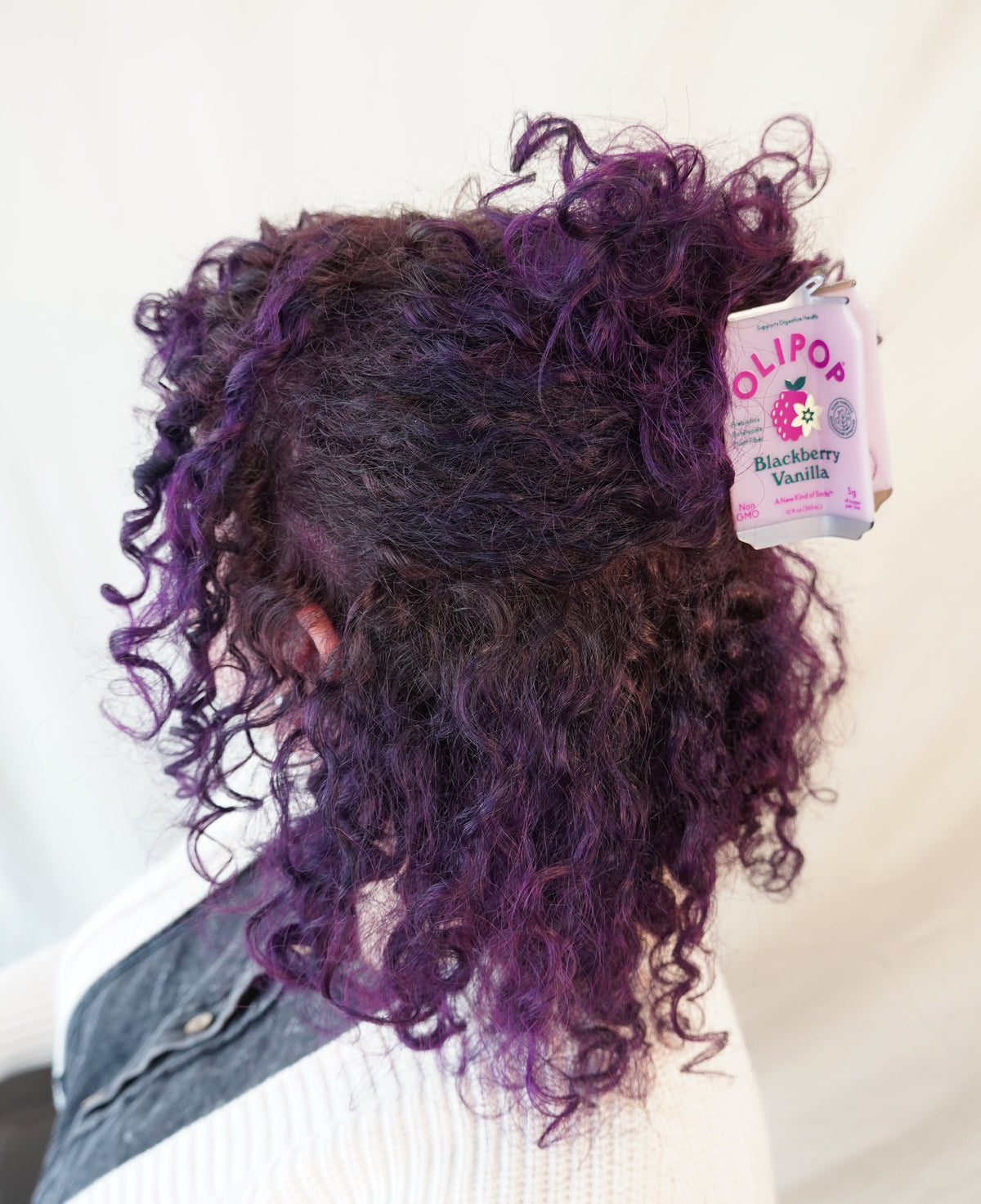 A person shown from the back wearing a white ribbed sweater and denim jacket, with curly hair in deep purple and black tones. A Olipop Blackberry Vanilla hair claw is holding their hair half up