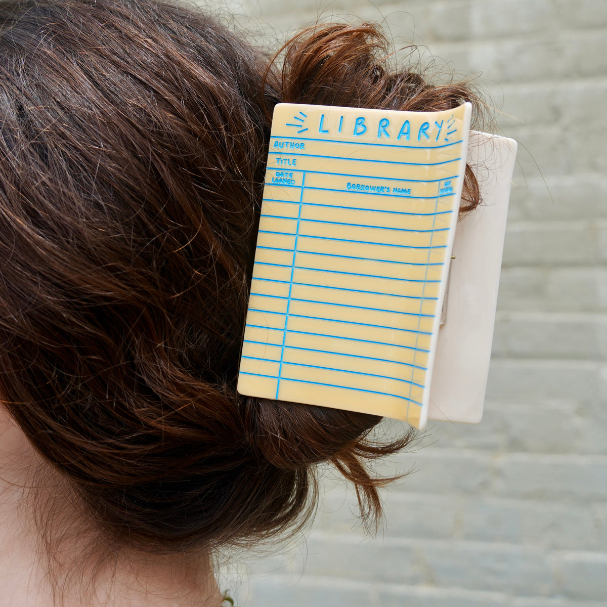 A close up of the Library Card Hair Claw in a person's brown hair, showing the yellow card with blue lines and 'LIBRARY' text.