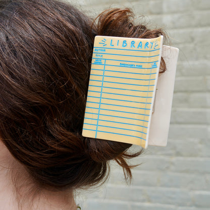 A close up of the Library Card Hair Claw in a person's brown hair, showing the yellow card with blue lines and 'LIBRARY' text.