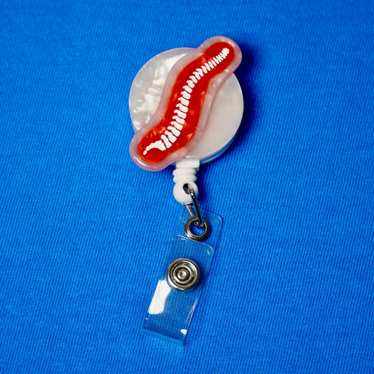 The Spine Badge Reel, a white pearl round reel with a red and white spine design on the front and a clear badge strap, on a blue fabric background.