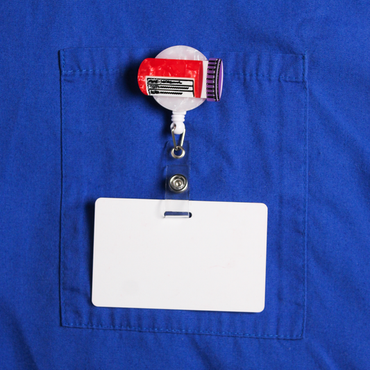 A badge reel on a blue fabric background, clipped to a pocket. The badge reel has a blood vial design on it, which is red with a purple cap. 