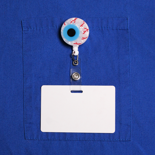 The Eyeball Badge Reel clipped to the pocket of a blue scrub top with a blank white ID card. The reel is a pearly white eyeball with red veins and a blue iris.