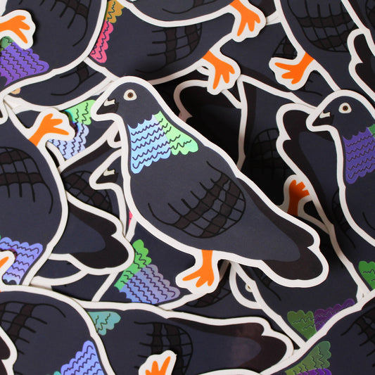 A pile of Pigeon Stickers, each a dark grey pigeon with orange feet and an iridescent green, blue and purple neck, with one sticker in focus on top.