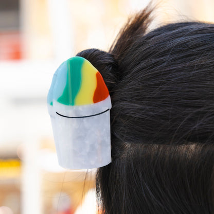 A person with dark hair wearing the Snowball Hair Claw, a pearly white cup topped with red, yellow, green and blue shaved ice, in a bun.
