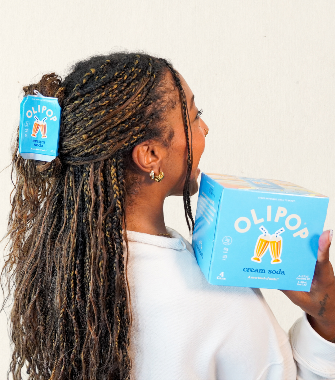 A woman with long braided hair is shown from the side, holding a blue OLIPOP cream soda box next to her head where a matching OLIPOP cream soda hair claw in her hair
