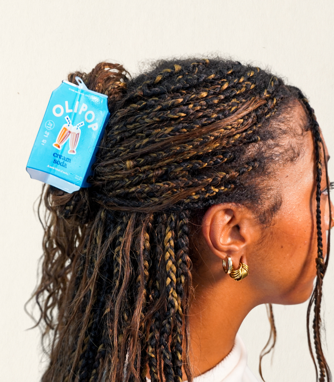 A close-up side profile of a person with long braided hair, wearing a Olipop cream soda hair claw in their hair, half up