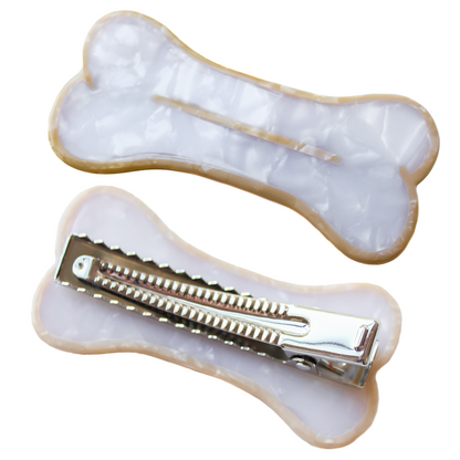 A hair clip shaped like a bone. The bone is white with a beige outline and line in the center. The image contains two bone clips, one flipped over showing the alligator clip mechanism