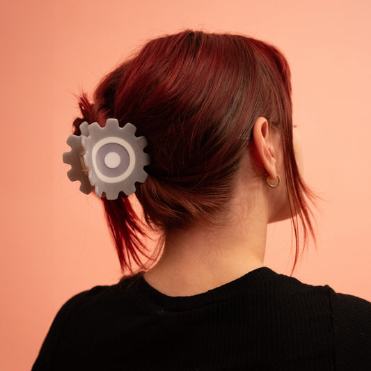 A person with red hair wearing the Engineer Gear Hair Claw, a grey cog shaped claw with white and grey rings in the centre, in a low ponytail against a peach background.