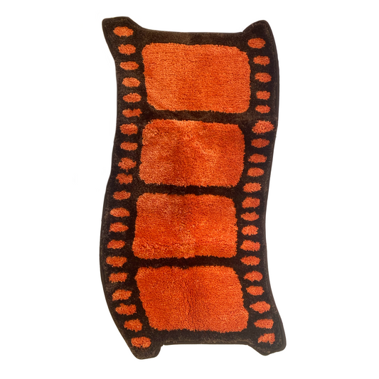 The Film Reel Rug on a white background, a wavy film strip shape in dark brown with four orange frames and orange sprocket holes along the edges.