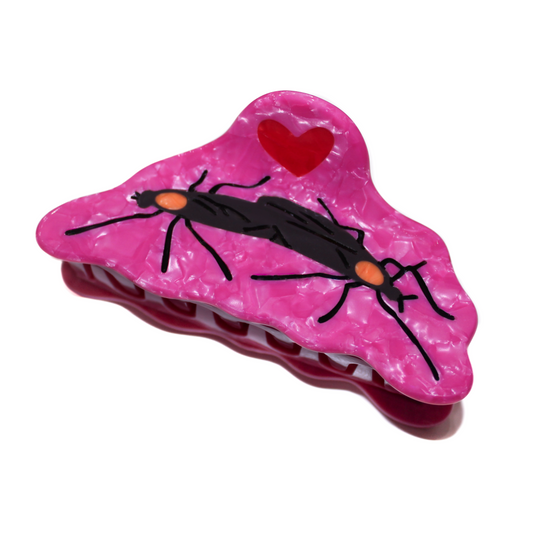 The Lovebug Fuggbug Claw, a hot pink pearl hair claw with a wavy edge, a pair of black lovebugs with orange heads and a red heart above them, on a white background.