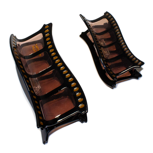A size comparison of the Film Reel Mega Claw next to the standard size Film Reel claw on a white background. Both are black and translucent brown, shaped like a strip of film with yellow sprocket holes.