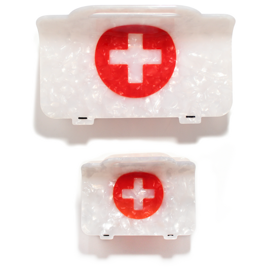A size comparison of the First Aid Mega Claw above the standard size First Aid Hair Claw, both pearly white first aid kits with a red circle and white cross, shown from the front on a white background.