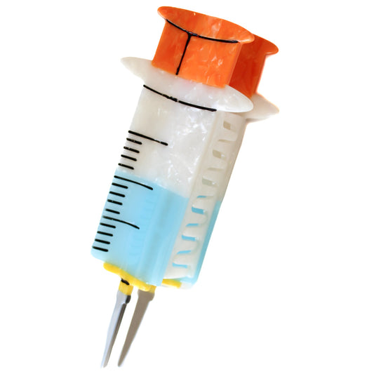 The Syringe Hair Claw, a hair claw designed to look like a syringe with an orange plunger, a white barrel with black measurement marks, blue liquid and a grey needle, on a white background.