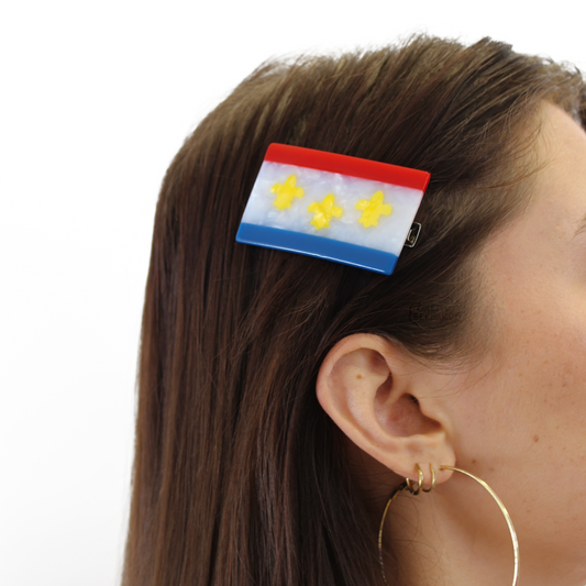 A person with brown hair and gold hoop earrings wearing the New Orleans Flag Hair Clip, a red, white pearl and blue flag with three yellow fleur-de-lis, on a white background.