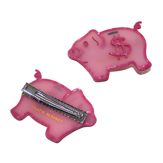 Two Piggybank Clips on a white background, one showing the front, a pink pig with a dollar sign and coin slot, the other flipped to show the silver alligator clip and gold 'COLETTE BERNARD' logo.