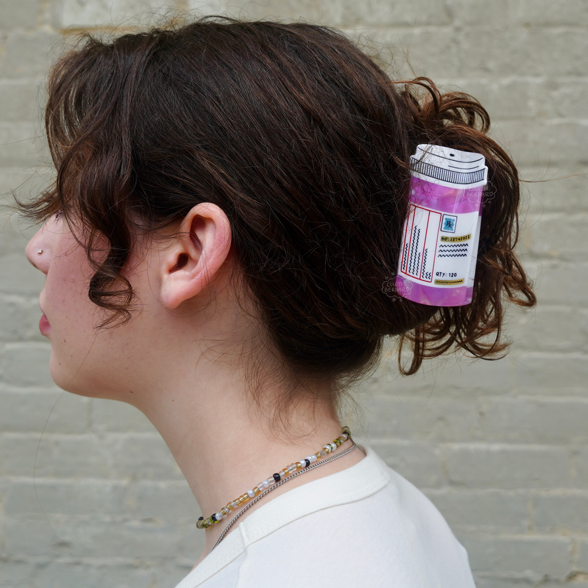 A person with short curly brown hair shown in profile wearing the Pink Prescription Bottle Hair Claw in a bun, against a pale green brick wall.