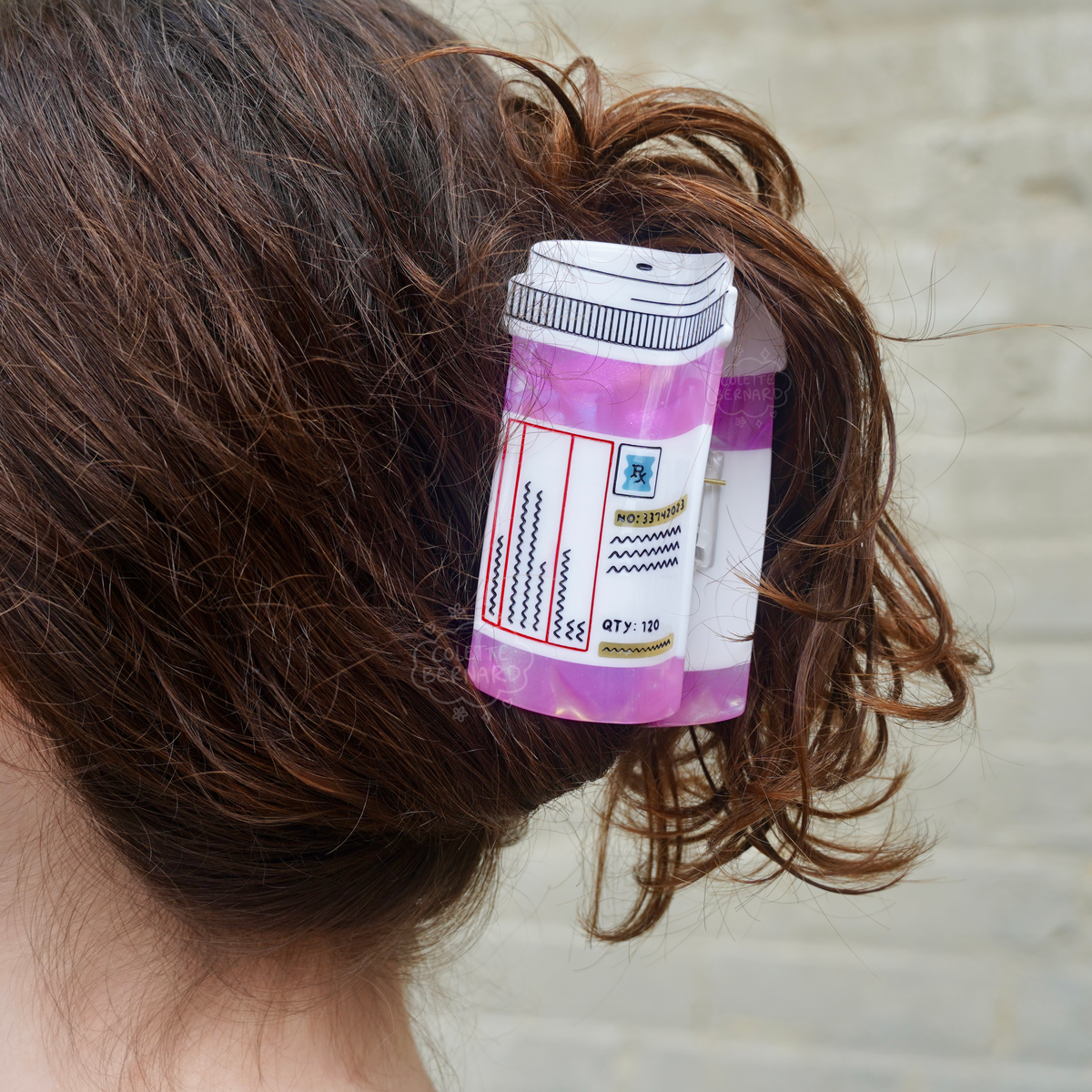 A close up of the Pink Prescription Bottle Hair Claw holding up a person's brown hair in a bun, against a pale green brick wall.