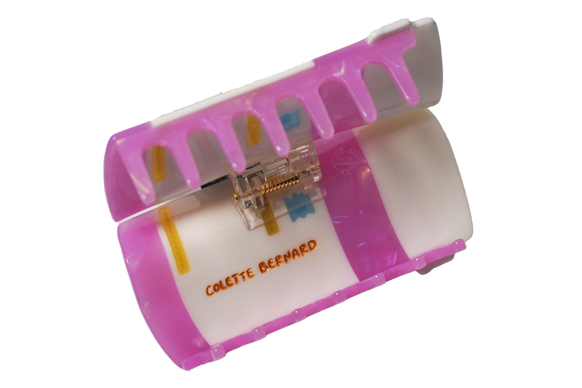 The Pink Prescription Bottle Hair Claw held open on a white background, showing the pink teeth, the spring mechanism and the orange Colette Bernard logo.