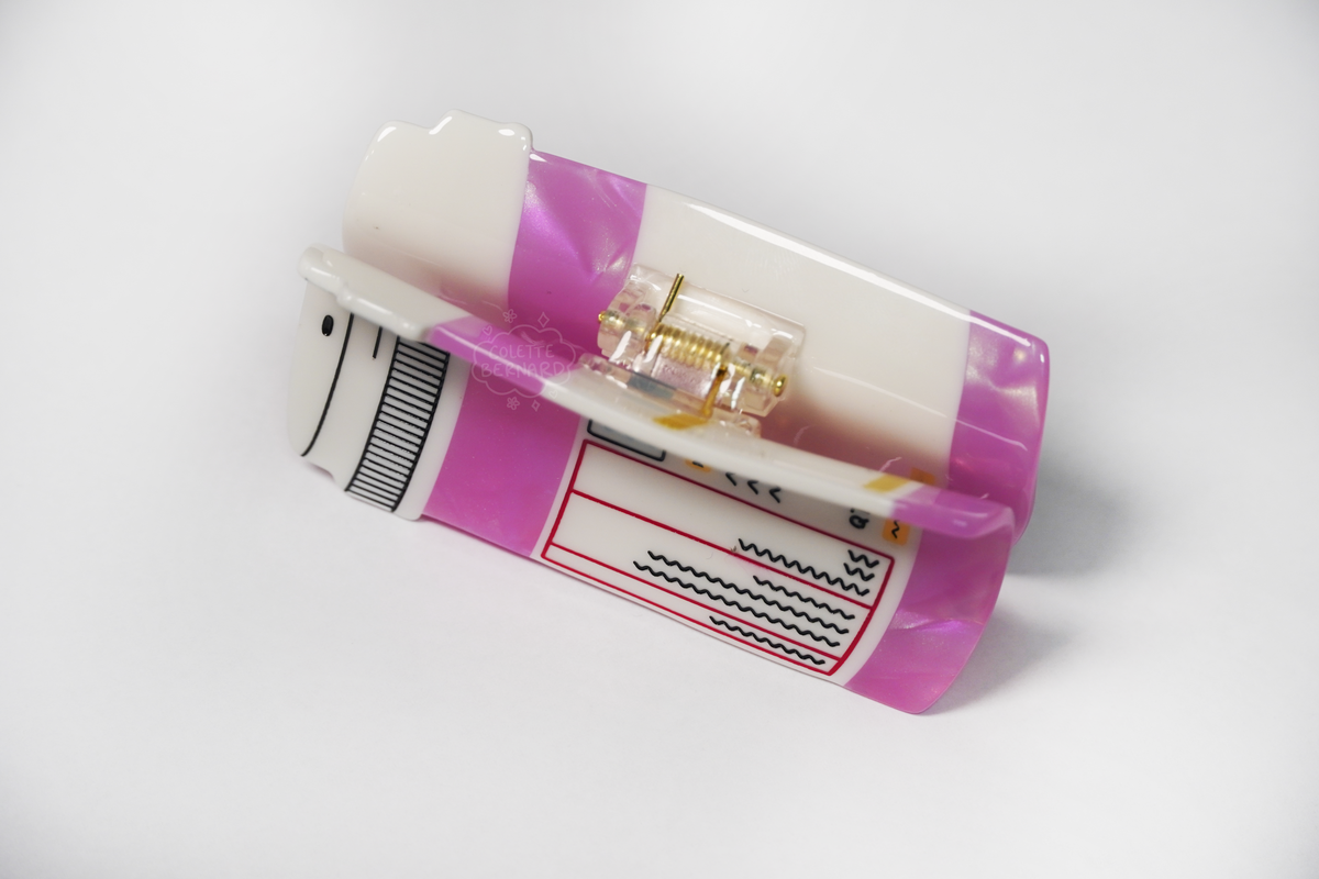 The Pink Prescription Bottle Hair Claw lying on its side on a white background, a top down view showing the spring mechanism, the white cap and the label.