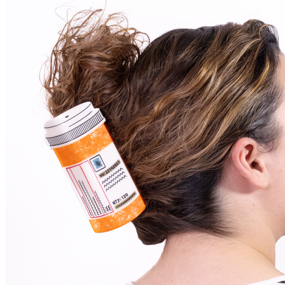 A person wearing the Prescription Bottle Mega Claw in a curly updo. The claw is orange with a white cap and a pharmacy label with an Rx symbol and 'QTY: 120'.