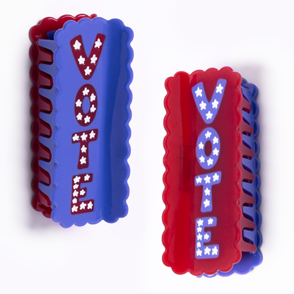 Two Vote Hair Claws on a white background, one blue with 'VOTE' in red star-filled letters and one red with 'VOTE' in blue star-filled letters, both with scalloped edges.